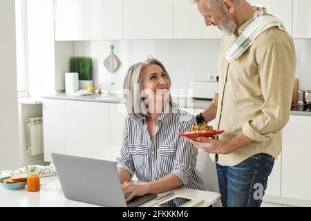 Senior husband preparing breakfast to happy wife working from home office. Stock Photo