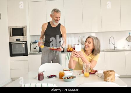 Happy older mature family couple having breakfast using phone in kitchen. Stock Photo