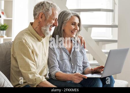 Happy older middle age couple using laptop looking at computer sitting on couch. Stock Photo