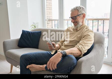Happy older middle aged man using phone relaxing sitting on couch at home. Stock Photo