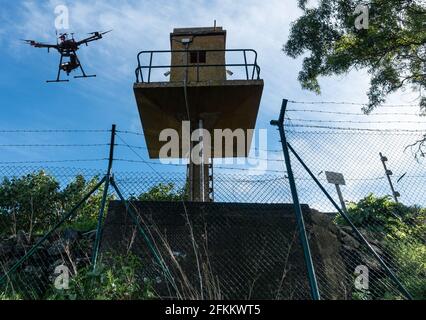 Drone flying above watchtower behind security, razor wire, barbed wire fence.