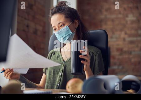Portrait of interior designer wearing protective mask holding a blueprint and thermos flask while working on new poject, sitting at the table in her Stock Photo