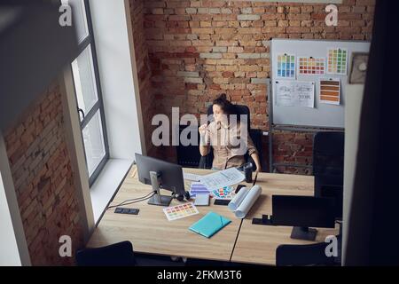 High angle view of thoughtful interior designer woman looking at the window, holding glasses while sitting in modern office Stock Photo
