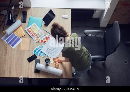 Top view of interior designer or architect in casual wear with messy hairdo working on a blueprint for new project, standing in her office on a Stock Photo
