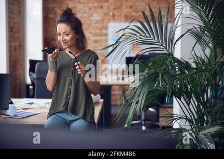 Cheerful interior designer woman holding glasses and smiling while making a call, standing in modern green office on a daytime and talking about new Stock Photo