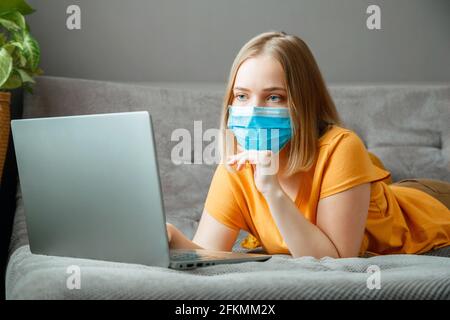 Woman in medical protective mask work using laptop at Home office lies on couch. Teen girl in mask Online learning education via laptop covid 19 Stock Photo