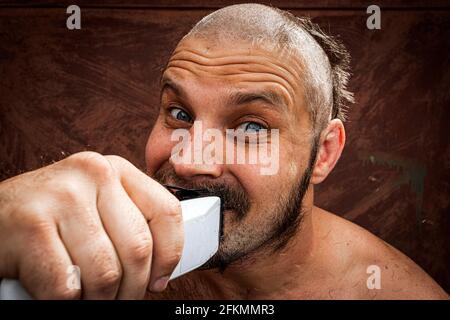 Caucasian man trying to shave with an electric razor. A brutal bald man holds a razor in his hand and shaves stubble on a metal background Stock Photo