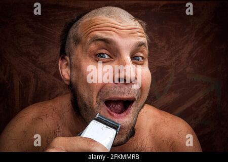 Caucasian man trying to shave with an electric razor. A brutal bald man holds a razor in his hand and shaves stubble on a metal background Stock Photo