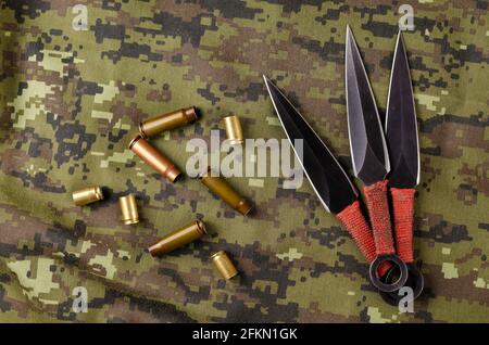 Black Throwing knives and casings on camouflage cloth. Three special knives with red handles crossed. Military theme. View from above at an angle. Cop Stock Photo