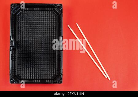 Chopsticks and Empty rectangular black plastic container for products on a red background. Plastic container for sushi set delivery. Take away lunch b Stock Photo