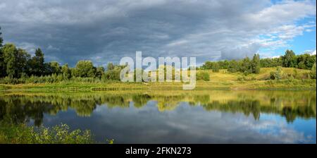 Stormy dramatic sky over the calm lake and green hills during sunset.Beautiful panoramic landscape.Beauty of evening nature. Stock Photo