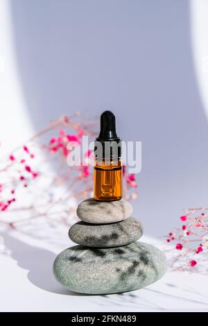 Small bottle of serum on stack of basalt stones on white background. Trendy shadows. Beauty pipette dropper on pyramid of pebbles with Gypsophila or b Stock Photo
