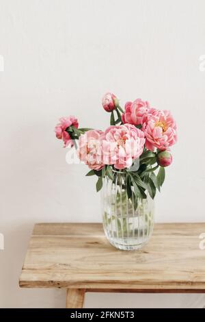 Floral still life scene. Pink peonies flowers, bouquet in glass vase on wooden table. White wall. Selective focus, blurred background. Wedding Stock Photo