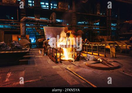 Foundry workshop interior, molten iron pouring from blast furnace into ladle container and workers founders control process, heavy metallurgy industry. Stock Photo
