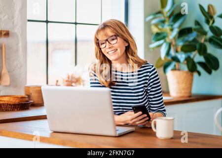 Happy middle aged woman using her laptop and having video call while sitting in the kitchen at home. Stock Photo