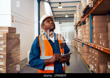 Young male factory worker maintaining stock records in large warehouse with stack of cardboard boxes Stock Photo