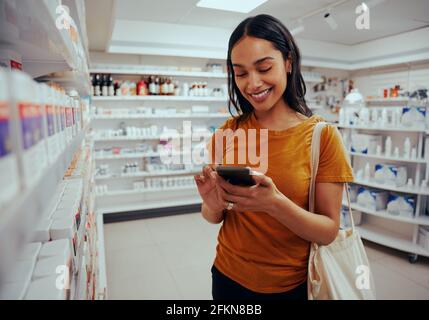 Young woman smiling while using smartphone standing against shelf in pharmacy Stock Photo