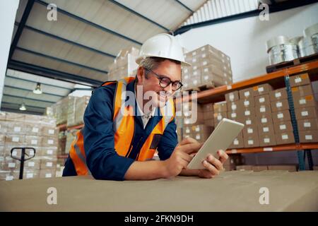Happy male industrial employee checking digital tablet wearing protective work wear at warehouse Stock Photo