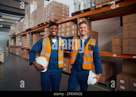 Smiling portrait of two multi ethnic warehouse workers holding safety hardhat in hand at factory Stock Photo