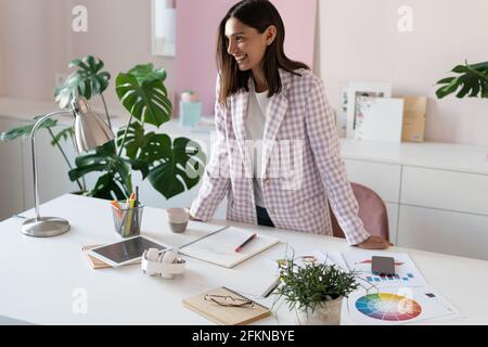 Confident young woman in smart casual wear smiling while standing near her working place Stock Photo