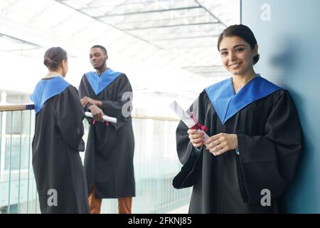 Group of young people wearing graduation gowns indoors in modern university interior, focus on smiling Middle-Eastern woman holding diploma and lookin Stock Photo