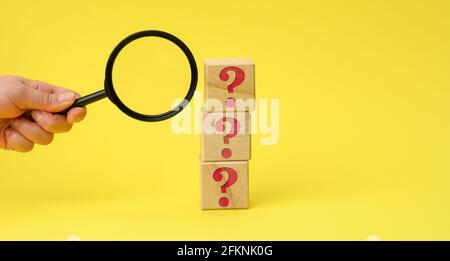 wooden cubes with question marks and a female hand holds a magnifying glass on a yellow background. The concept of finding answers to unknown question Stock Photo