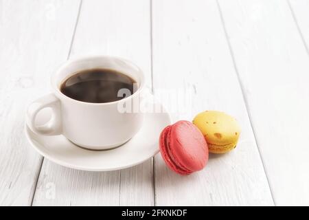 Cup of hot steaming coffe and two macaroons on a white wooden table. Sweet breakfast with delicious almond meringue cookies. High key image. Stock Photo