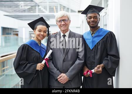 Waist up portrait of two college graduates holding diplomas while posing with mature professor and smiling at camera Stock Photo