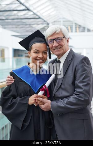 Vertical waist up portrait of young African-American woman holding diploma while posing with mature professor and smiling at camera Stock Photo