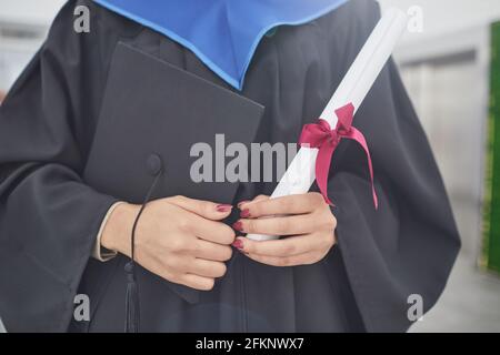 Cropped portrait of unrecognizable young woman holding diploma and graduation cap while standing outdoors during ceremony, copy space Stock Photo