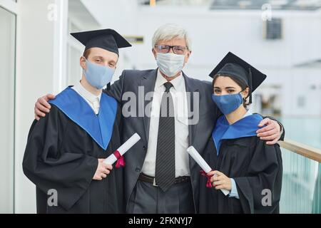 Waist up portrait of two college graduates wearing masks while posing with mature professor indoors Stock Photo