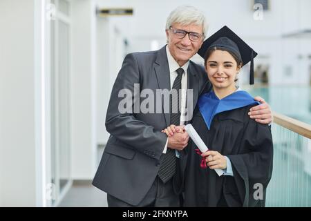 Waist up portrait of smiling young woman posing with father during graduation ceremony indoors Stock Photo