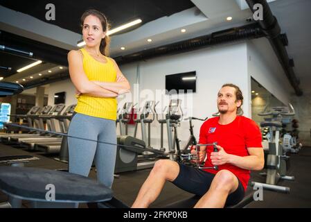 Young athletic Caucasian woman standing next to young man exercising on a rowing machine in the modern gym Stock Photo