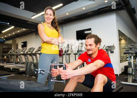 A smiling young athletic Caucasian woman standing next to young man while having fun and exercising on a rowing machine in the modern gym Stock Photo