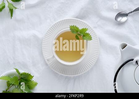 Nettle tea cup, fresh stinging nettle herbs. Flat lay, top view, off white textile tablecloth. White ceramic teapot. Stock Photo