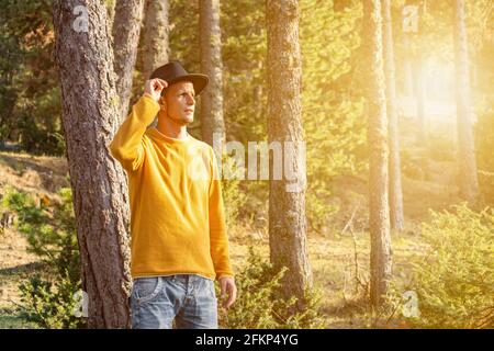 Young stylish man in hat in the forest.Portrait tourist cowboy in yellow sweater in woodland at sunset .Beautiful landscape with Sunlight and copy spa Stock Photo