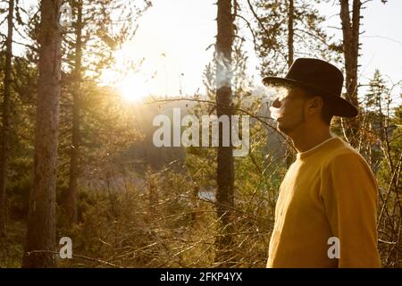 Young stylish man smoking in hat in the forest.Portrait tourist cowboy with smoke in yellow sweater in woodland at sunset .Beautiful landscape with Su Stock Photo