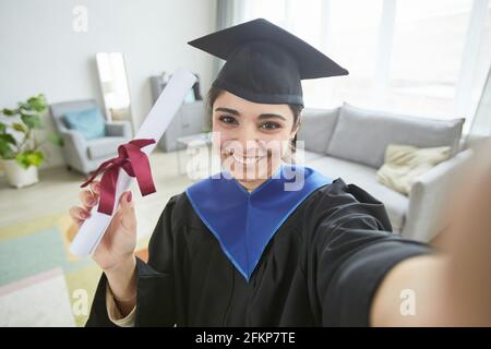 Smiling young woman wearing graduation gown while taking selfie at home or video blogging Stock Photo