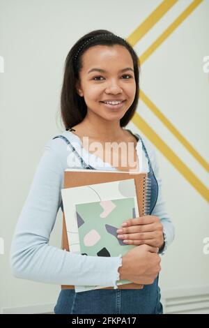 Vertical portrait of mixed-raced young woman smiling at camera in school while standing against wall and holding notebooks Stock Photo