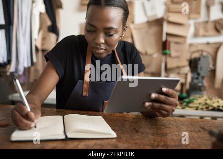 African female leather worker using a tablet and writing notes Stock Photo