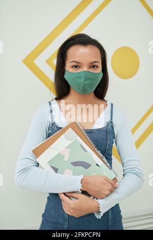 Vertical portrait of mixed-raced young woman wearing mask in school while standing against wall and holding notebooks Stock Photo