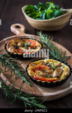 Two vegetarian tartlets on a wooden board and a wooden bowl with fresh field salad on a rustic table, vertical stock photo Stock Photo