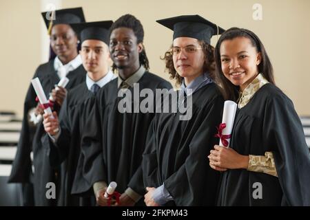 Multi-ethnic group of happy young people wearing graduation robes standing in row and looking at camera Stock Photo