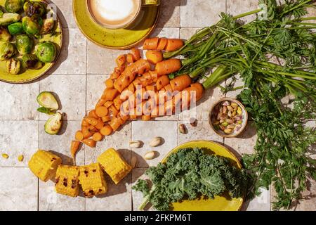 Bunch of grilled carrot roots with green leaves, Brussels sprouts on green craft plate, salad leaves, nuts, apricot jam grilled corn and a cup of coff Stock Photo