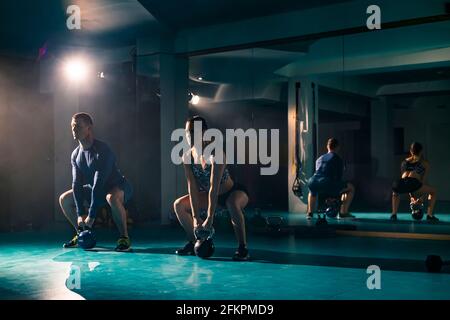 Woman and a man simultaneously doing a sumo high pull with kettle-bell exercise at the gym. Stock Photo