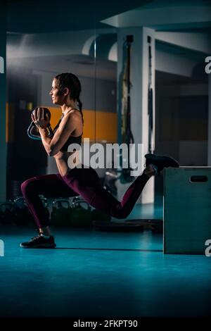 A young woman doing a split squats exercise in the empty gym with weights and other exercise equipment in her surroundings. Stock Photo