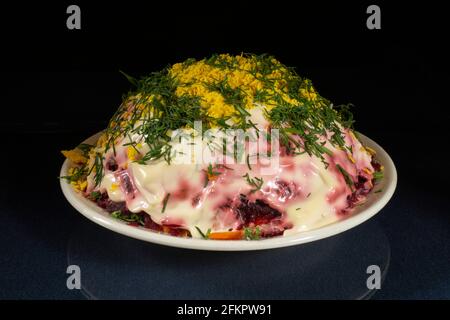 Herring under a fur coat with mayonnaise in a white plate on the table with a reflection. Food for the holiday on a black background Stock Photo
