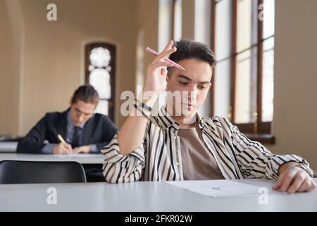 Portrait of young teenage boy taking exam at desk in school auditorium and thinking, copy space Stock Photo
