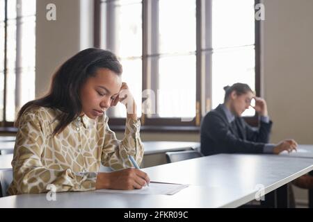 Side view portrait of two students taking exam in row while sitting at desk with social distancing, focus on young African-American woman in foregroun Stock Photo
