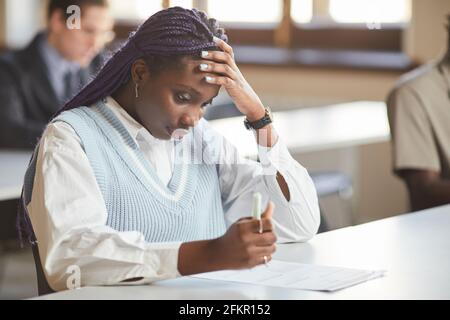 Portrait of young African-American woman taking exam in school while sitting at desk in college and thinking, copy space Stock Photo